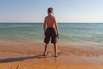A teenage boy on the beach in a mask with a snorkel and fins wants to swim underwater in the sea.