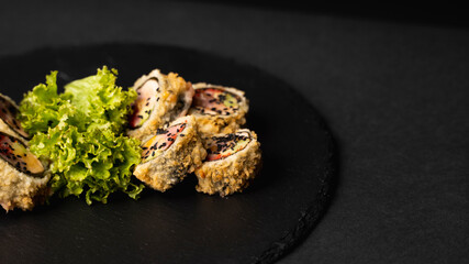 Custom sushi roll in tempura with nori, fresh salmon, tuna, avocado, masago caviar, drizzled with pineapple sauce with salad pouring as decoration on a black plate on a black table and background.
