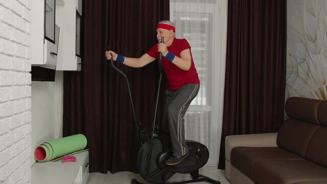 Senior elderly caucasian man in sportswear using orbitrek in living room doing sport training warming up cardio exercises at home. Active old grandfather during coronavirus lockdown. Healthy lifestyle