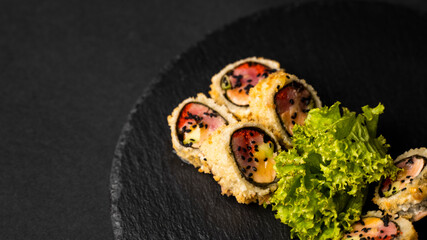 Custom sushi roll in tempura with nori, fresh salmon, tuna, avocado, masago caviar, drizzled with pineapple sauce with salad pouring as decoration on a black plate on a black table and background.