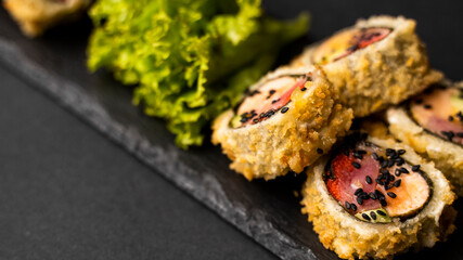 Custom sushi roll in tempura with nori, fresh salmon, tuna, avocado, masago caviar, drizzled with pineapple sauce with salad pouring as decoration on a black plate on a black table and background.