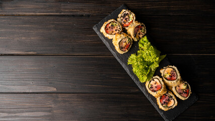 Fototapeta na wymiar Custom sushi roll in tempura with nori, fresh salmon, tuna, avocado, masago caviar, drizzled with pineapple sauce with salad pouring as decoration on a black plate on a wooden table and background.