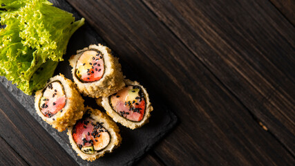 Custom sushi roll in tempura with nori, fresh salmon, tuna, avocado, masago caviar, drizzled with pineapple sauce with salad pouring as decoration on a black plate on a wooden table and background.