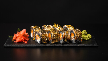 Custom sushi roll with nori, cuttlefish ink, fresh salmon, cream cheese, pepper pumpkin, eel, unagi sauce and edible gold leaf with red ginger, wasabi on a square plate on black table and background.