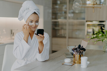 Pleased young woman wears bathrobe and towel wrapped on head cares for under eye skin applies beauty patches looks at herself in mirror sits at table with laptop and cosmetic tools poses at home