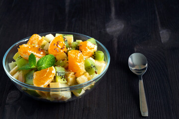 Refreshing fruit salad with spoon. healthy salad from fruit with green mint on a wood table. Mixed fresh banana, orange, kiwi, fruit on the black background. copy space.