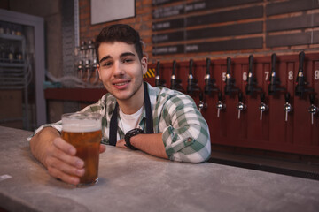 Young bartender smiling joyfully, serving glass ov delicious light beer, copy space