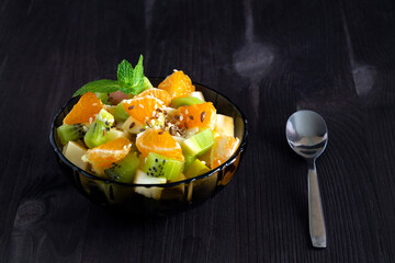 healthy salad from fruit with green mint on a wood table. Mixed fresh banana, orange, kiwi, fruit on the black background. copy space. Refreshing fruit salad with spoon.