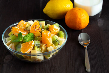 fruit salad in glass bowl. Refreshing fruit salad with lemon. healthy salad with green mint on a wood table. Mixed fresh banana, orange, kiwi, fruit with glass of milk on the black background.