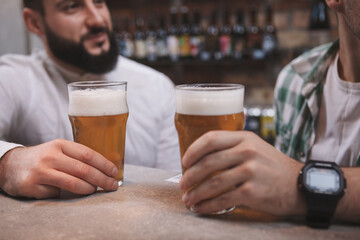Cropped close up of beer glasses male friends are holding while chatting at the bar