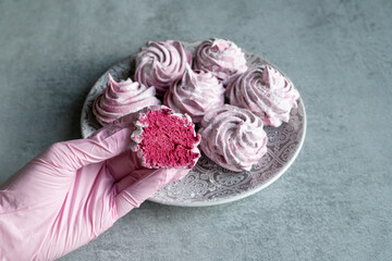 A pink-gloved hand holds a pink marshmallow with marshmallows in the background. Delicious sweets, dessert, healthy sweets