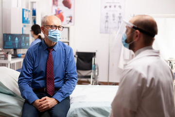 Senior man talking with doctor about treatment sitting on bed waring face mask as safety precaution Global health crisis, medical system during coronavirus pandemic, sick elderly patient in private
