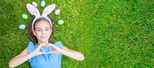 Smiling young girl wearing bunny ears on Easter day lying on green summer grass and showing heart sign. Top down view. Empty space for text