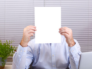 A man in an office at a computer holds a vertical sign in his hands and covers his face.