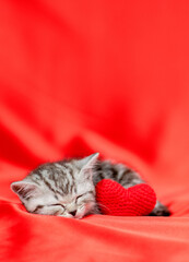 Tabby kitten sleeps with red heart on satin bedding. Valentines day concept. Empty space for text © Ermolaev Alexandr