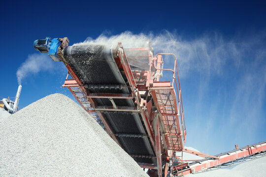Conveyor belt of a working mobile crusher, close-up, with blown away by the wind white stone dust against a blue sky.