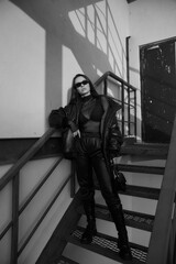 Stylish fashionable woman with makeup and long straight brunette hair in black leather bomber, joggers, high boots, bra, net body and sunglasses standing at red urban metal stairs