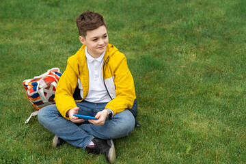Schoolboy sits on the lawn with  smart phone in his hands. Modern teenager in yellow jacket