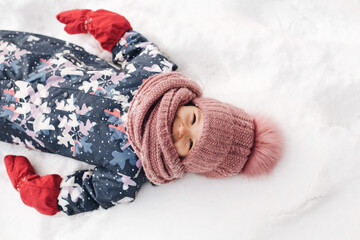 A little cute girl 3 years old in a winter overalls, a hat and a scarf lies on the snow in a snowdrift and smiles 