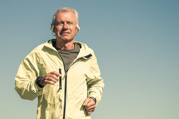 Happy excited senior man in wireless headphones jogging outside. Low angle, blue clear sky in...