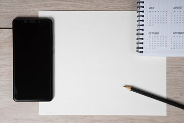 Top view of workplace. Empty paper blank, spiral calendar, black pencil and phone on wooden table.