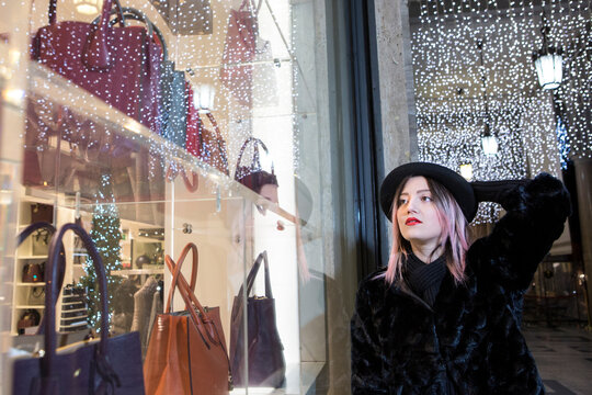 Bella ragazza con i capelli castani dipinti di viola con un cappotto e  un cappello nero  v&agrave; in giro per la citt&agrave; piena di illuminare  notturne a fare shopping sotto i portici