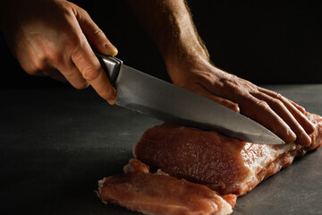 Cooking. Male hands with a knife cut a large piece of raw fresh meat on a stone gray background.