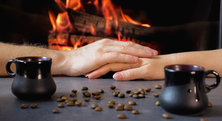 Fototapeta na wymiar Romantic coffee break. A couple drinks coffee from ceramic coffee cups against a backdrop of burning fire and coffee beans. Close-up on hands.