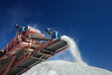 Conveyor belt of a working mobile crusher machine, close-up, with blown away by the wind white stone dust against a blue sky. - obrazy, fototapety, plakaty