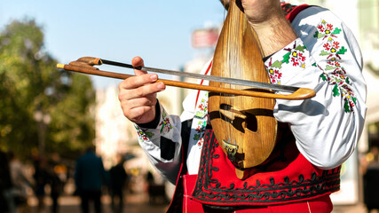 Bulgarian folk musician - violinist in traditional national costume plays an old stringed instrument - gadulka on street of Plovdiv city, Bulgaria. Bulgarian folklore and culture. Street performer - obrazy, fototapety, plakaty