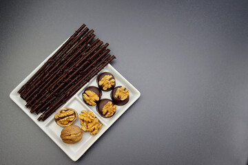 Long chocolate sticks, chocolate candies and peeled walnuts in a white square plate on a gray background. Chocolate and walnuts.