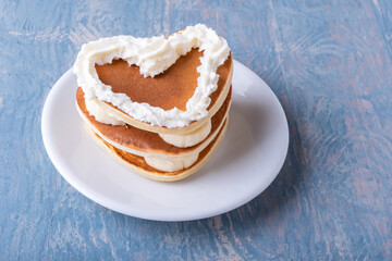 Valentine's day creative breakfast. Homemade heart shaped pancake with banana decorated with white cream on a white plate on a blue wooden table, copy space, top view, close-up.