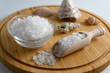 Little wooden scoop and glass bowl full of sea salt on wooden cutting board on white background