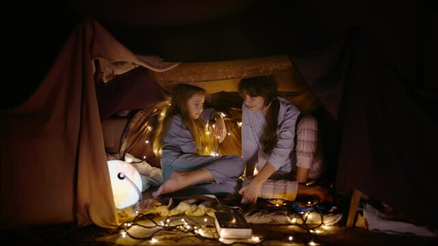 Children play in tent made of blankets and pillows in home. Two little girl friends play together in secret place late evening