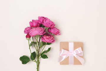 Gift box with a tied bow and bouquet of rose flowers on a beige background. Minimal festive composition with copyspace.