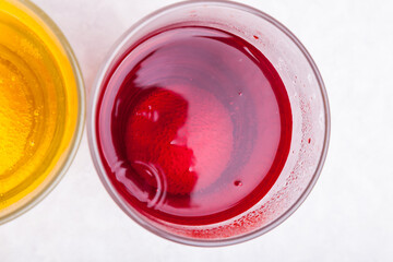 Top view of a glass with bright red liquid close-up - juice with a dye and air bubbles on a white background next to a glass with yellow liquid. Unhealthy foods and unhealthy drinks high in sugar.