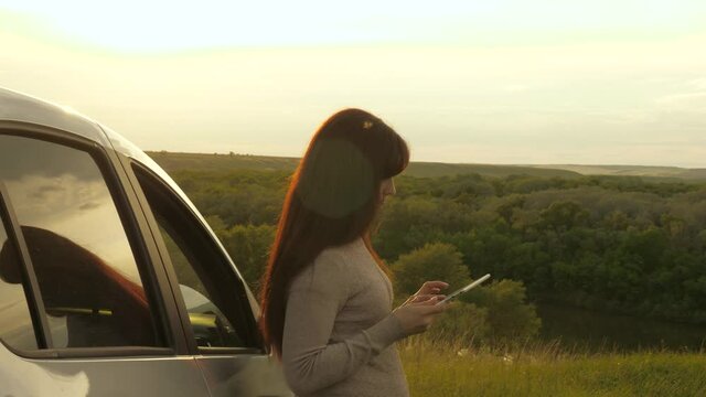 A free woman is resting next to the car, checking route in navigator. A tourist with a smartphone stopped at campsite by car, looking at sunset. Tourist driver holding modern gadget in his hands.