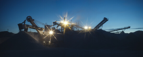 Silhouette of excavator and of stone-crushing quarry equipment in the light of spotlights at night, industrial panorama with a spectacular starry cross filter.