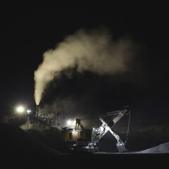 Excavator on the background of rock stone crushing equipment in the light of spotlights at night, industrial panorama.