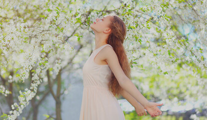 Happy young woman raising her hands up in a spring blooming garden on a flowers background