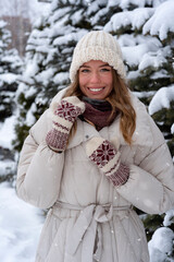 Beautiful smiling young woman in a hat and warm clothes in winter. Portrait of a cheerful girl in a snow forest.