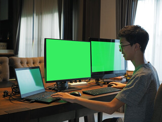 Rear view of an Asian boy wearing glasses working on three computer screens at home. He is smiling and laughing while working at home. The screens are with green screen.
