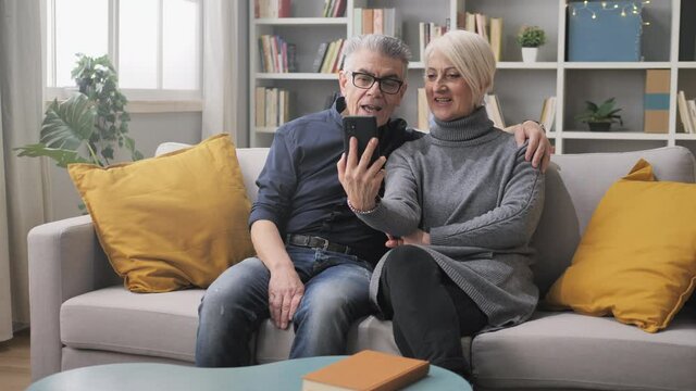 Happy elderly couple sit on sofa make a video call conference using smart phone at home,old caucasian man and woman holding smartphone have a funny conversation with friends