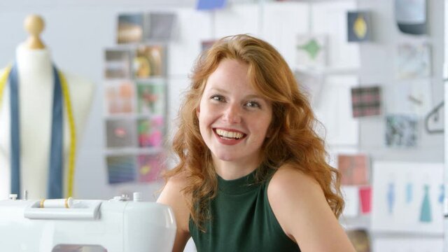 Camera pans across fashion studio as female clothes designer works at sewing machine and smiling at camera - shot in slow motion