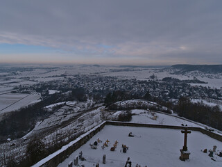 Beautiful winter landscape with panoramic view over village Wurmlingen in Baden-Württemberg, Germany surrounded by snow-covered fields and forests on cloudy day with small cemetery in front.