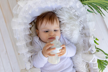 a small baby girl lies in a beautiful cradle in a white bodysuit at home and holds a bottle of milk...