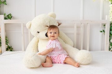 happy little baby girl six months old sitting on a white bed in pink clothes, with a big Teddy bear and smiling