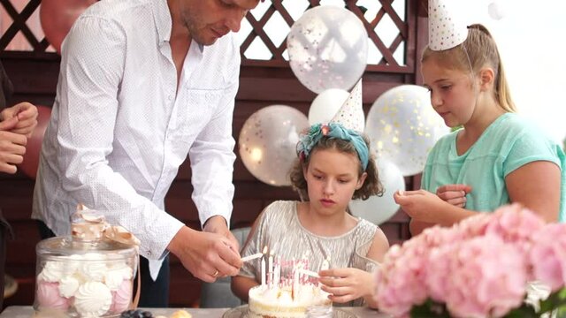 Family Celebration Or A Garden Party Outside In The Backyard. The Birthday Girl, Parents And Guests Light Candles On The Birthday Cake. Children's Birthday