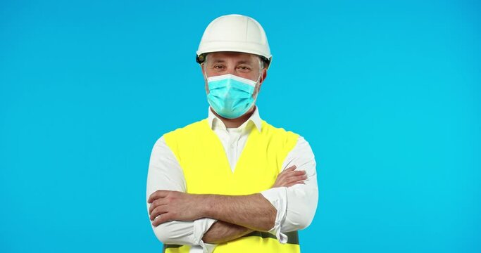 Portrait of happy joyful Caucasian adult handsome professional male builder engineer wearing medical mask and helmet standing isolated on blue background in studio looking at camera. Worker concept