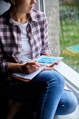 Obraz premium Woman sitting at the window, writing in notebook plan, to do list after quarantine end, coronovirus ending